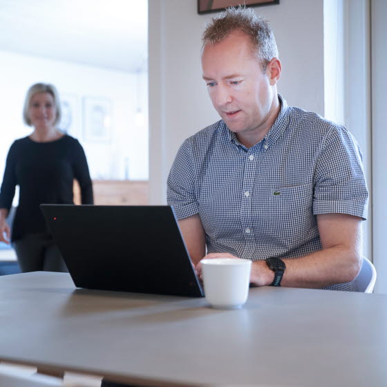 Mand sidder ved spisebord med computer og kvinde kommer gående i baggrunden