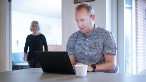 Mand sidder ved spisebord med computer og kvinde kommer gående i baggrunden