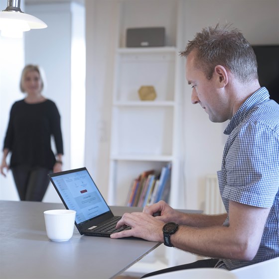 Mand arbejder på PC ved spisebord mens kvinde kommer gående ind
