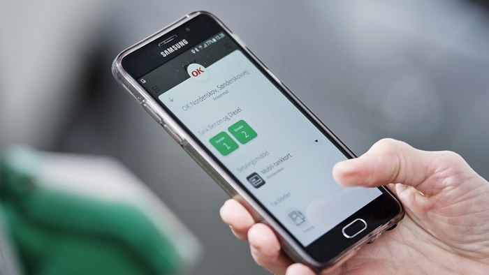 Mobiltelefon med OK's app på skærmen og tankpistol i baggrunden