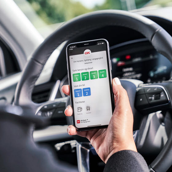 Hånd holder mobil med OK's app, sidder i bil er ved at parkere