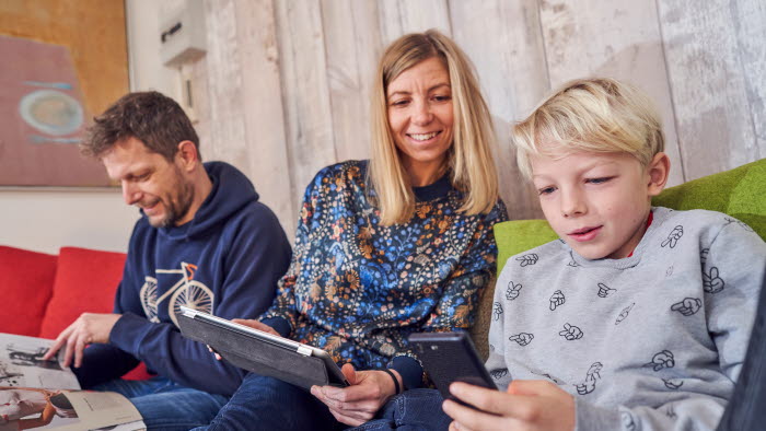 Forældre og søn sidder i sofa sammen med tablet og mobiltelefon foran sig