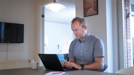 Mand sidder med bærbar computer ved spisebord med TV på væggen i baggrunden