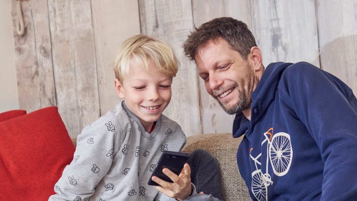 Dreng og far sidder i sofa og smiler mens de kigger på mobiltelefon