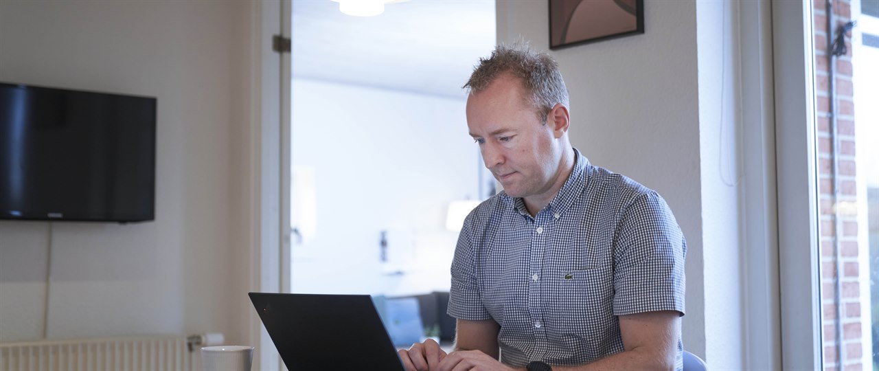 Mand sidder med bærbar computer ved spisebord med TV på væggen i baggrunden