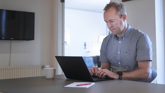 Mand sidder med bærbar computer ved spisebord med TV på væggen i baggrunden