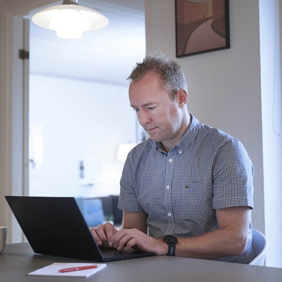 Mand sidder med bærbar computer ved spisebord med TV på væggen i baggrunden