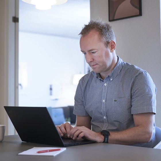 Mand sidder med bærbar computer ved spisebord med TV på væggen i baggrunden