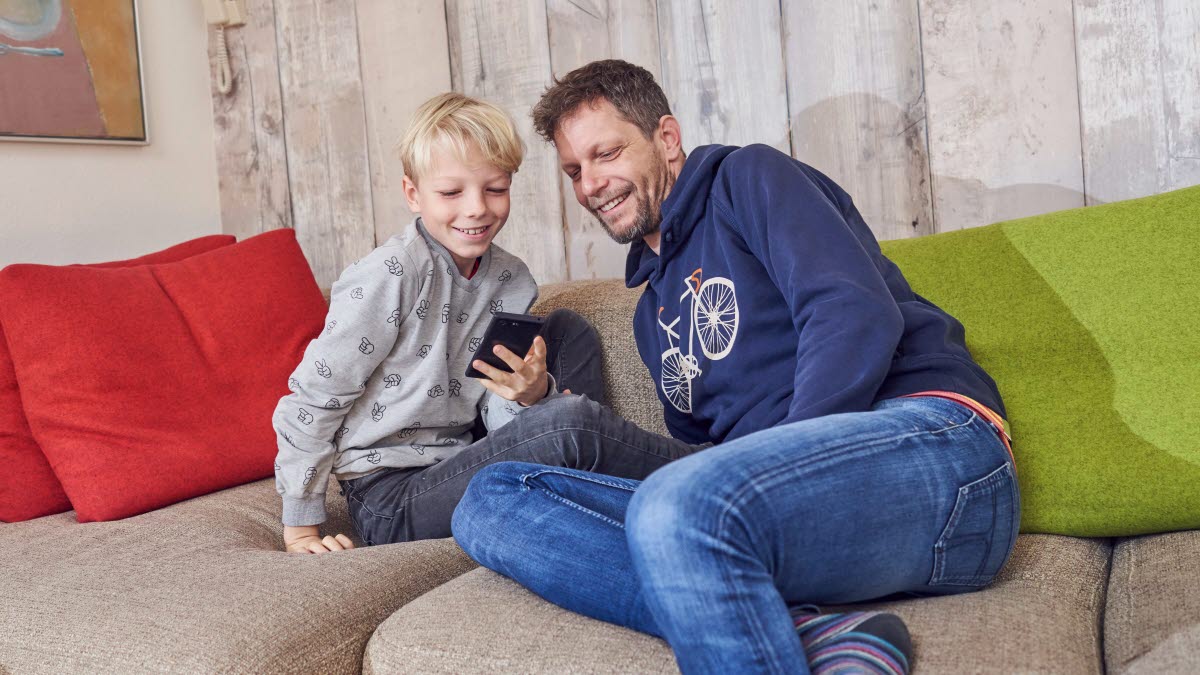 Dreng og far sidder i sofa og smiler mens de kigger på mobiltelefon