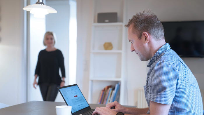 Mand arbejder på PC ved spisebord mens kvinde kommer gående ind