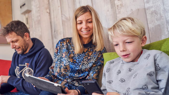 Forældre og søn sidder i sofa sammen med tablet og mobiltelefon foran sig